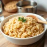 A close-up of fluffy, perfectly cooked brown rice served in a white bowl, garnished with parsley, with a wooden spoon on the side and uncooked rice in the background