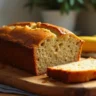 A freshly baked loaf of honey banana bread with a golden crust, sliced on a wooden board, with honey and ripe bananas beside it.