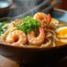 A steaming bowl of shrimp ramen noodles with soft-boiled egg, green onions, and sesame seeds, served in a ceramic bowl.