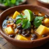 A bowl of Brazilian Mounjaro, featuring cassava, black beans, and coconut milk, garnished with cilantro and lime, served in a rustic clay bowl with spices and rice on the side.