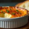 A creamy Crab Brûlée in a white ramekin with a golden caramelized sugar crust, garnished with chives and served with toasted baguette slices.