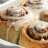 Ultra-realistic close-up of gooey chai-spiced cinnamon rolls with creamy frosting served in a white dish, captured with soft, natural lighting.