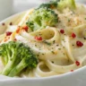A close-up shot of creamy garlic broccoli pasta with red pepper flakes, served in a white ceramic dish. The dish is coated in a rich garlic cream sauce, with vibrant green broccoli florets and a sprinkle of red pepper flakes for a spicy kick.