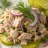Close-up of Dill Pickle Tuna Salad served on a plain white plate, showcasing creamy tuna, dill pickles, and fresh herbs with soft natural lighting