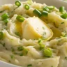 A close-up of a white dish filled with creamy Colcannon, featuring mashed potatoes blended with sautéed cabbage and green onions, topped with a melting pat of butter.