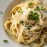 Close-up of Fettuccine Alfredo served in a plain white plate with rich creamy textures, garnished with Parmesan and parsley, captured in natural light.