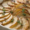 A top-down view of Crispy Balsamic Thyme Potato Torte in a white ceramic dish, featuring golden-brown, thinly layered potatoes arranged in a circular pattern with balsamic glaze and fresh thyme.