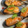 Japanese Dynamite Mussels topped with creamy Sriracha sauce, garnished with green onions, sesame seeds, and chili oil, served on a white plate