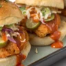 Close-up of three crispy bang bang chicken sliders with toasted buns, creamy spicy sauce, lettuce, pickles, and cilantro on a rustic wooden board.