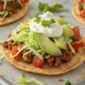 Close-up of crispy Mini Sheet Pan Tostadas topped with beans, cheese, avocado, and cilantro on a white plate