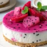 Close-up of no bake dragon fruit mini cheesecakes in glass jars topped with fresh dragon fruit cubes and mint leaves on a rustic wooden table