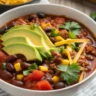 Close-up of a bowl of Easy Taco Soup topped with cheese, avocado, tortilla chips, and cilantro in a white ceramic bowl on a rustic table