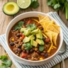 Close-up of a bowl of Easy Taco Soup topped with cheese, avocado, tortilla chips, and cilantro in a white ceramic bowl on a rustic table