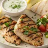 Grilled Greek Chicken with golden char marks served with tzatziki, lemon wedges, cucumber salad, and pita on a white plate