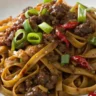 A close-up image of Mongolian Ground Beef Noodles served in a white plate with caramelized ground beef, green onions, and glossy soy sauce