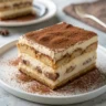 Close up photo of a costco tiramisu slice on a white plate showing creamy layers and cocoa topping.