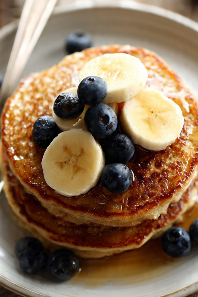 Stack of fluffy banana oat pancakes topped with fresh banana slices and blueberries on a white plate