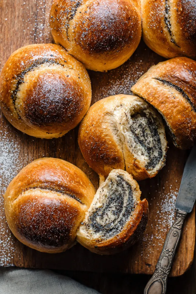 Freshly baked poppy seed buns with golden brown tops arranged on wooden board, one sliced open showing dark poppy seed filling inside