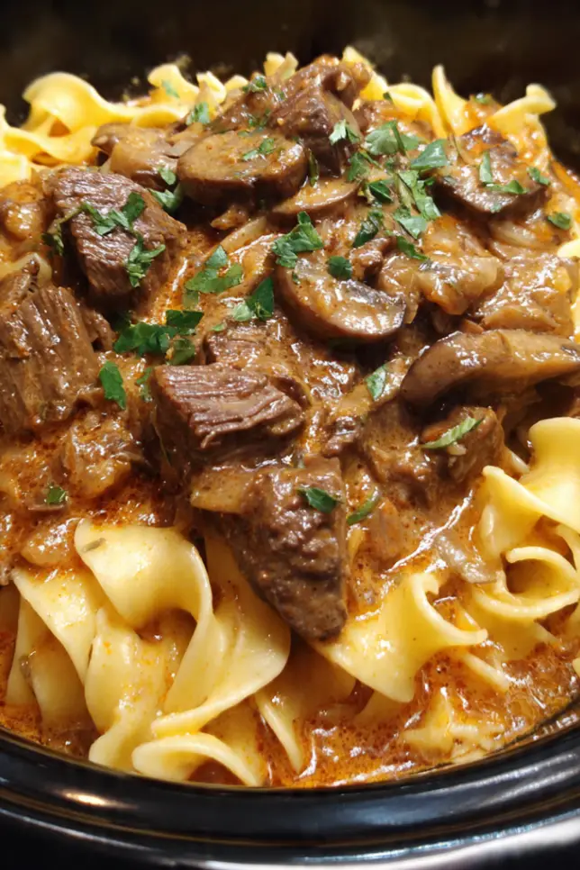 Homemade crockpot beef and noodles with tender shredded beef in rich gravy served in white bowl with fresh parsley garnish