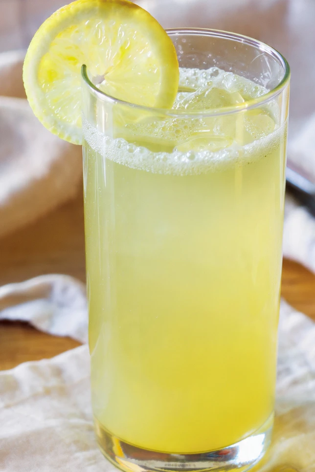 Tall glass of homemade German lemonade with ice cubes, lemon slice, and fresh mint garnish on wooden table with natural lighting