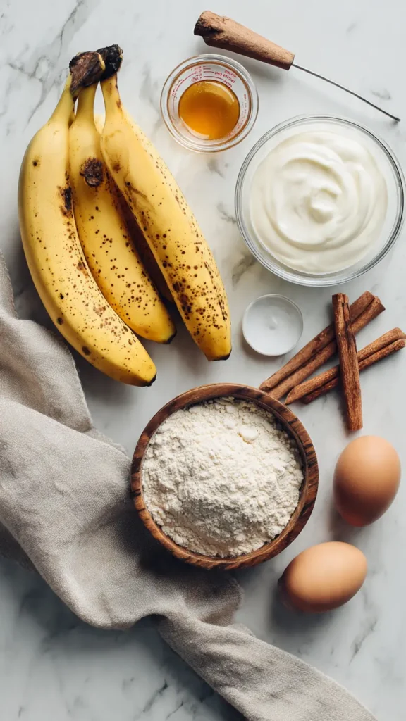 Overhead view of healthy banana cake ingredients including ripe bananas, maple syrup, Greek yogurt, eggs, and whole wheat flour on marble countertop