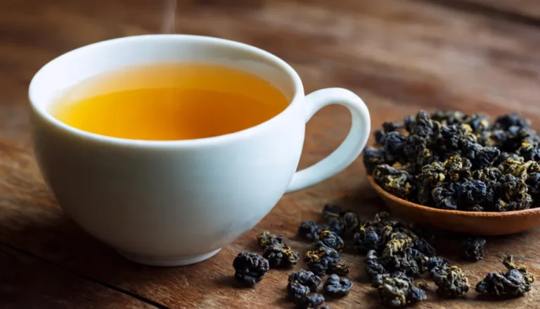 A steaming cup of amber oolong tea in a white ceramic cup with loose tea leaves on a wooden table