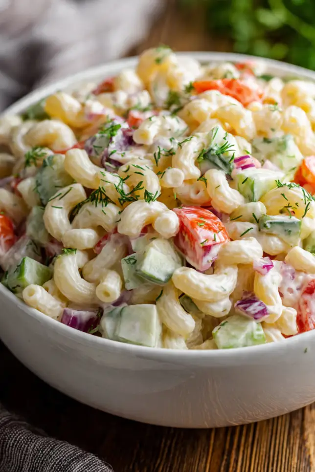 Classic macaroni salad in white bowl with creamy dressing, colorful vegetables, and fresh herbs on wooden table