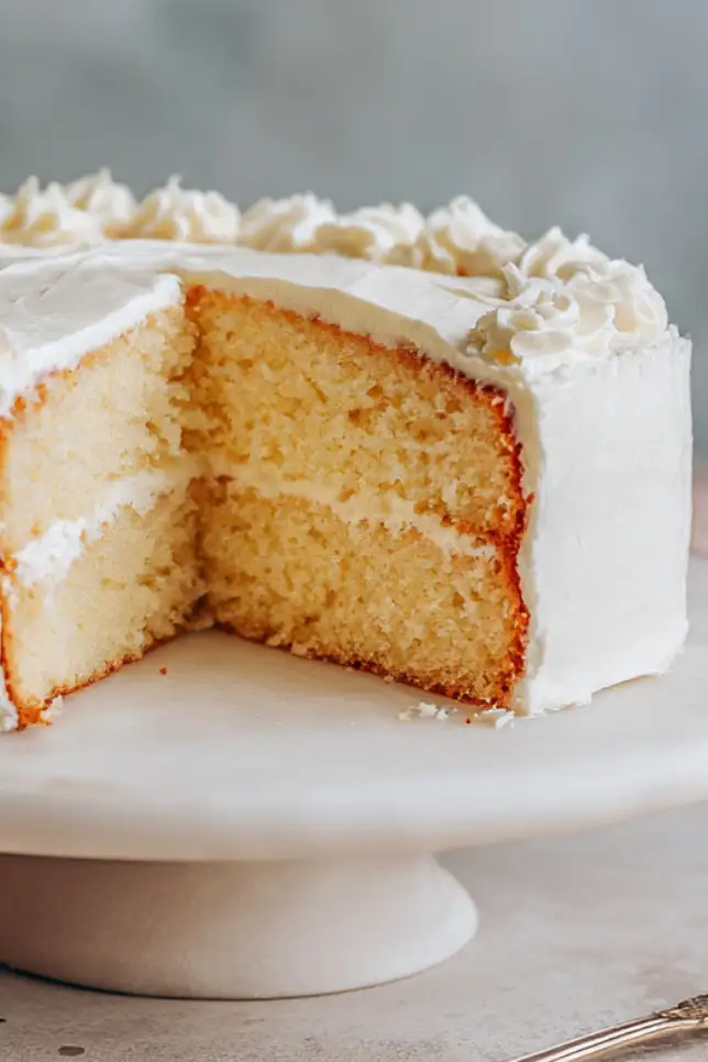 Homemade fluffy vanilla cake with white buttercream frosting on marble stand with slice removed showing light airy texture