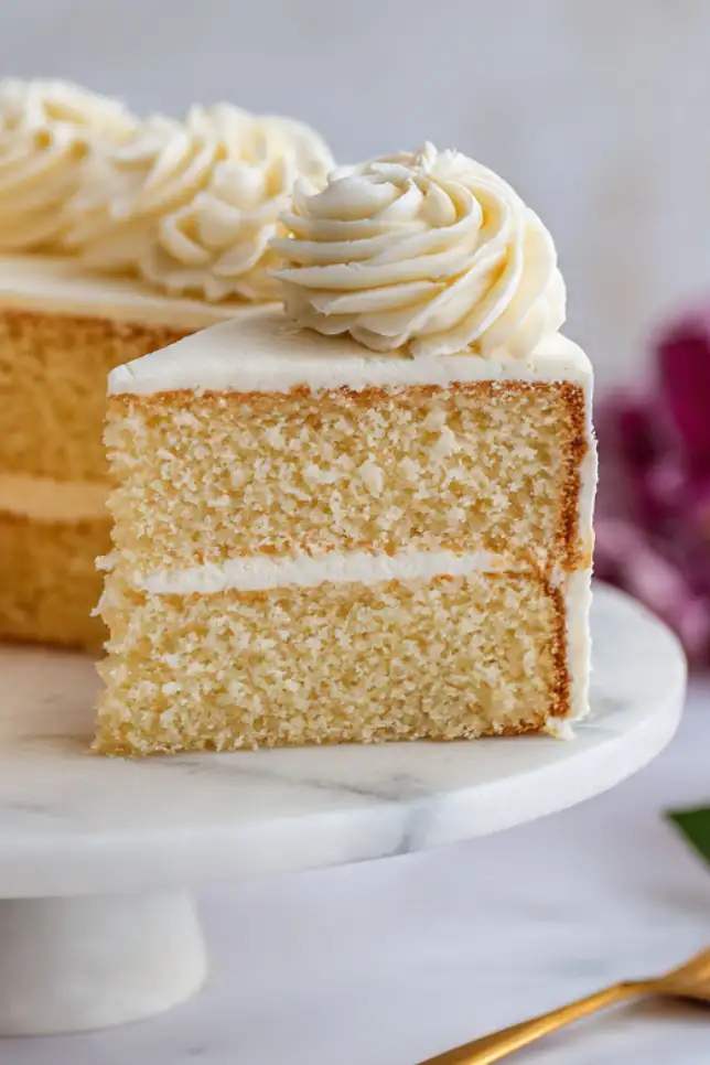 Homemade fluffy vanilla cake with white buttercream frosting on marble stand with slice removed showing light airy texture