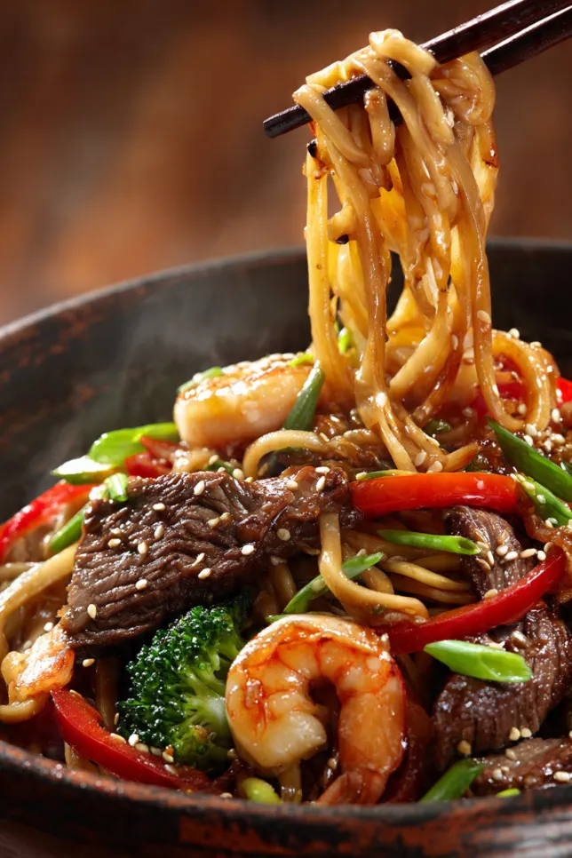 Homemade steak and shrimp stir-fry noodles in a white bowl with colorful vegetables and sesame garnish on a wooden table