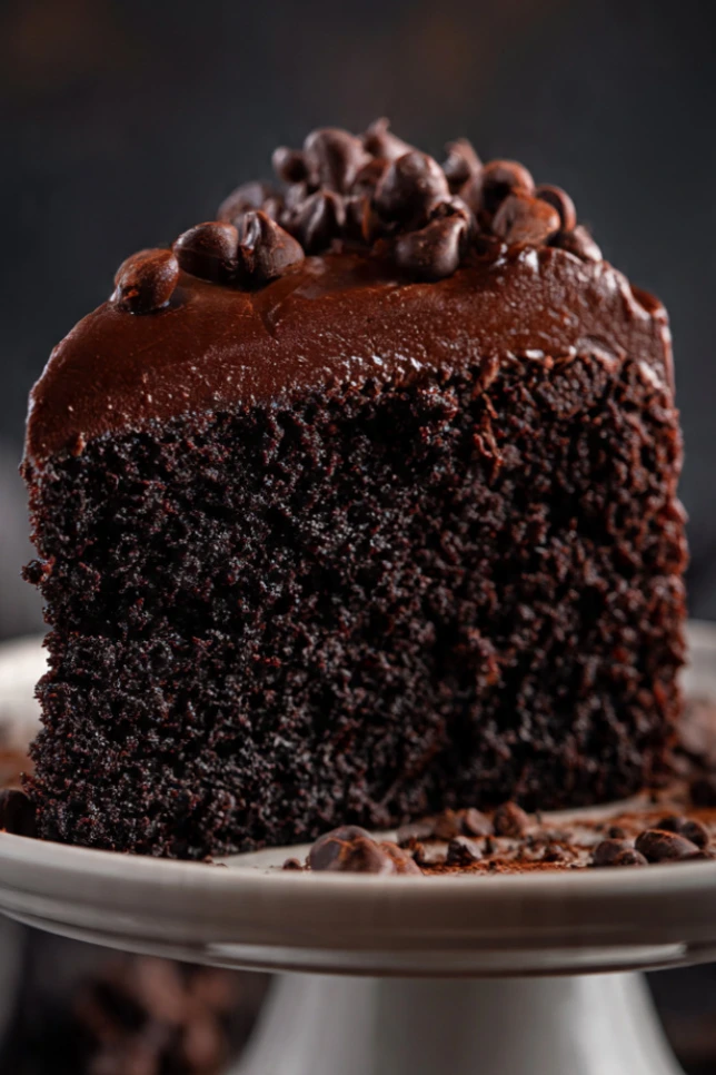 Slice of dense fudgy death by chocolate cake on white cake stand with chocolate chips scattered around