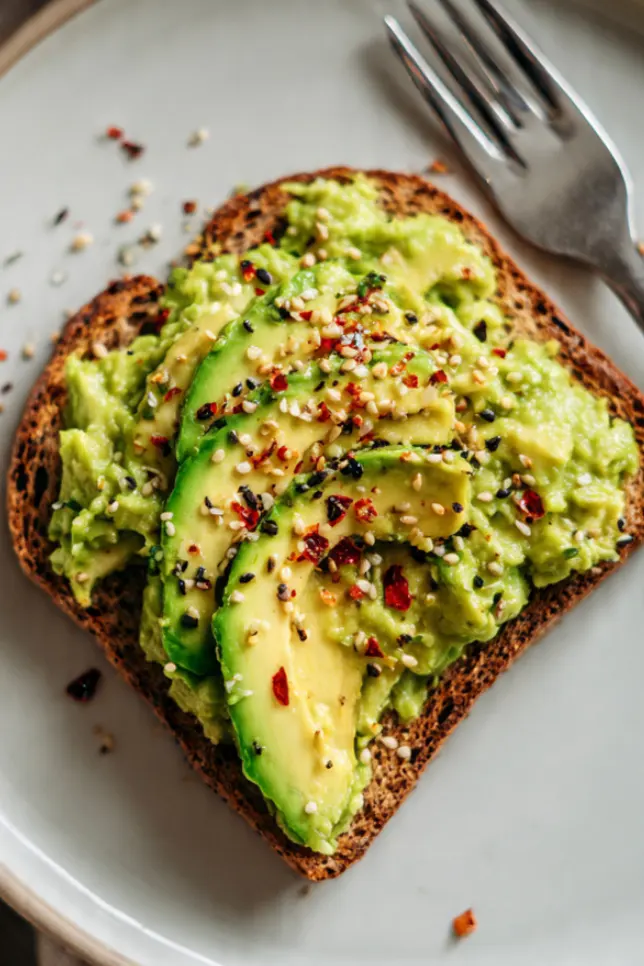 Flat lay of avocado toast ingredients including whole grain bread, avocado, lemon, red pepper flakes, and everything bagel seasoning