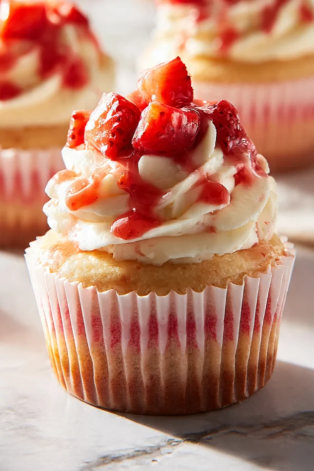 Homemade strawberry cheesecake cupcakes with cream cheese frosting and fresh strawberries on top, arranged on white marble surface