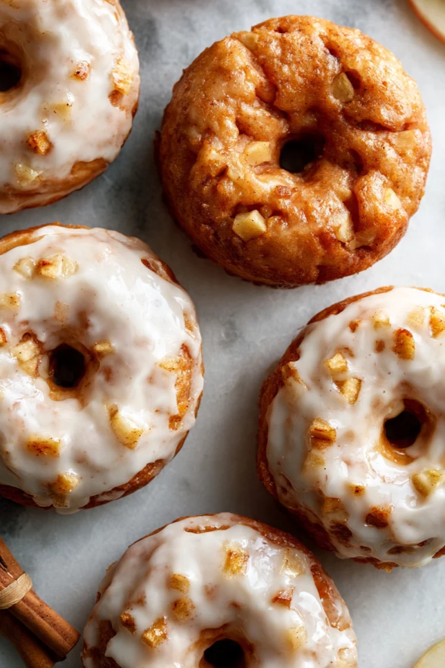 Baked apple fritter donuts with thick vanilla glaze on a marble surface surrounded by fresh apple slices and cinnamon sticks