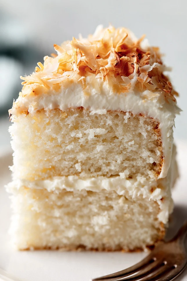 Tall fluffy coconut cake covered in cream cheese frosting and toasted coconut flakes on a white cake stand with one slice pulled away