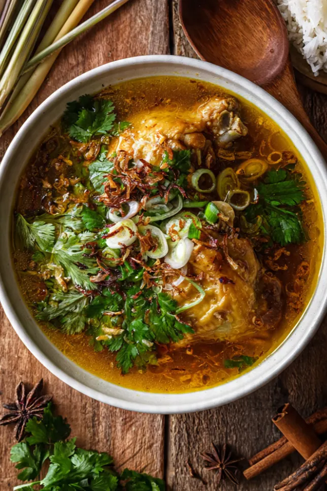 A steaming bowl of homemade Malay chicken soup (Sup Ayam) garnished with cilantro, fried shallots, and green onions, served with steamed rice on a wooden table