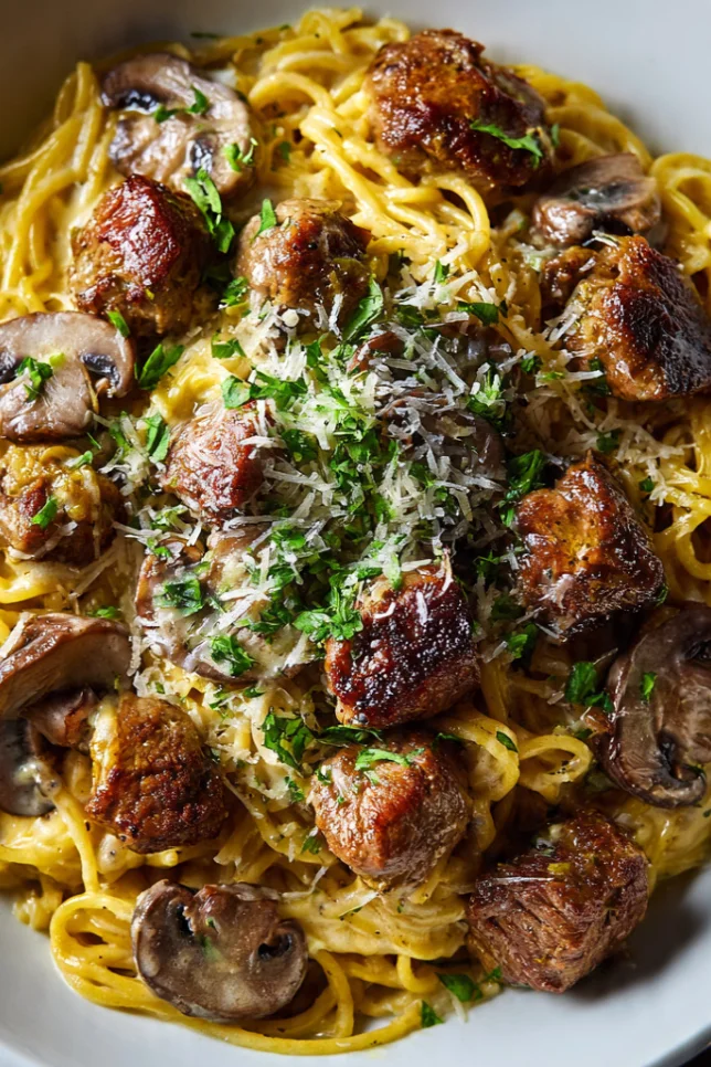Overhead view of creamy garlic steak bites spaghetti with mushrooms in a white bowl topped with parmesan and fresh parsley